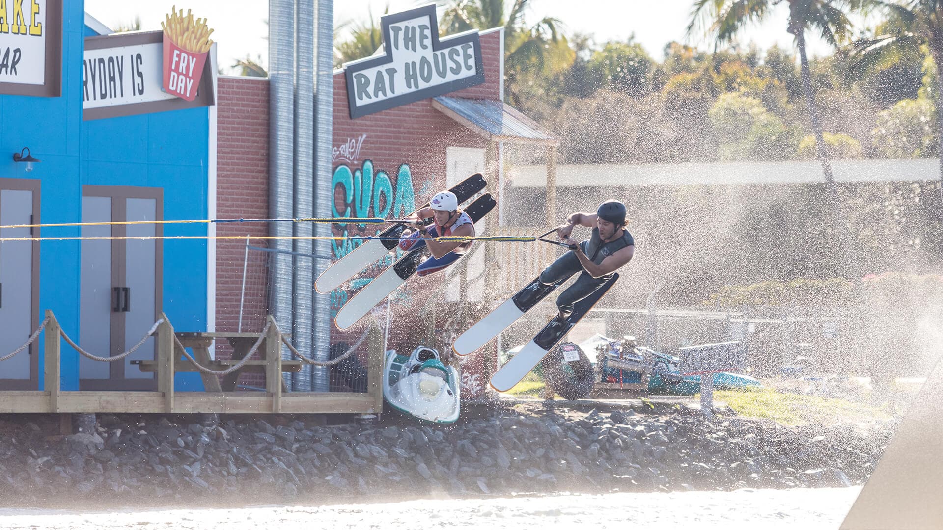 Two athletes performing a side-by-side jump on water skis at a cable ski park, wearing helmets and life vests, with colourful buildings and spray from the lake in the background.